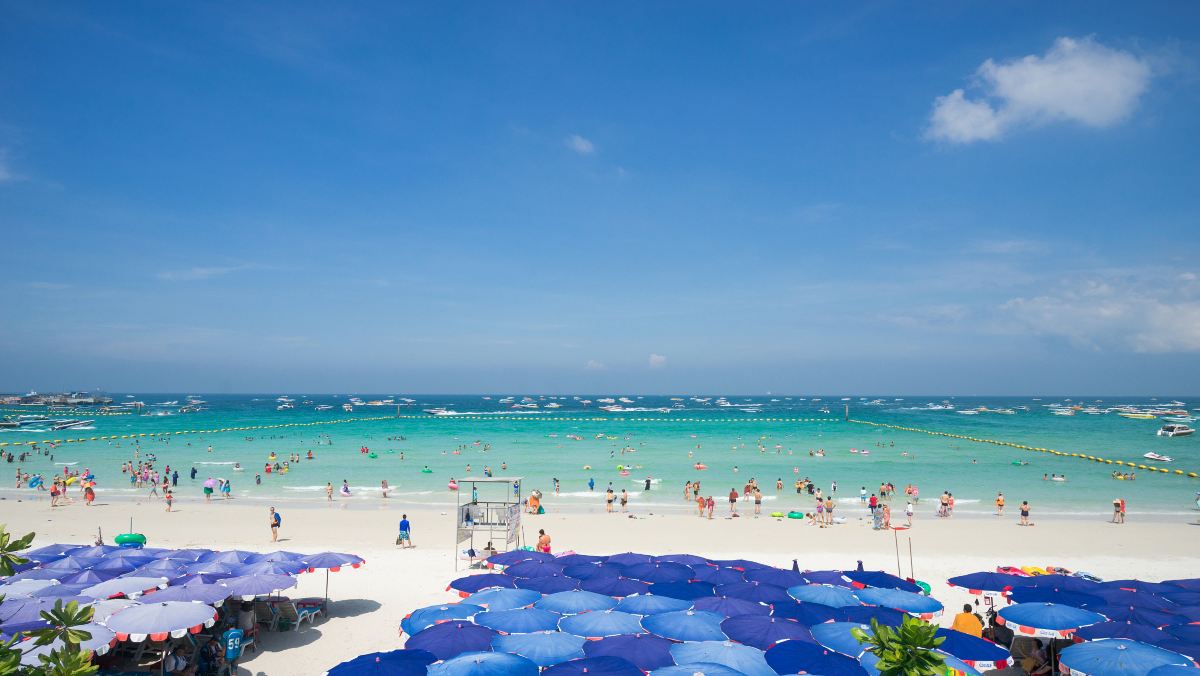 หาดพัทยาช่วงกลางวัน มีร่มชายหาดสีน้ำเงินและสปีดโบ๊ทจอดเรียงราย