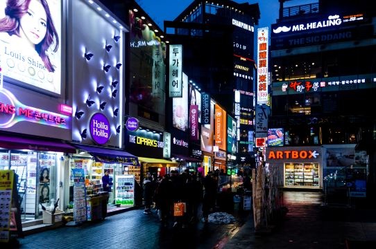 Neon shop signs over the pedestrian lanes of Myeong-dong in Seoul at night, with shoppers on the street