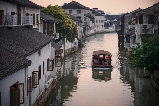 a boat traveling down a river next to a row of houses Via Unsplash (by 一只猫的橘)
