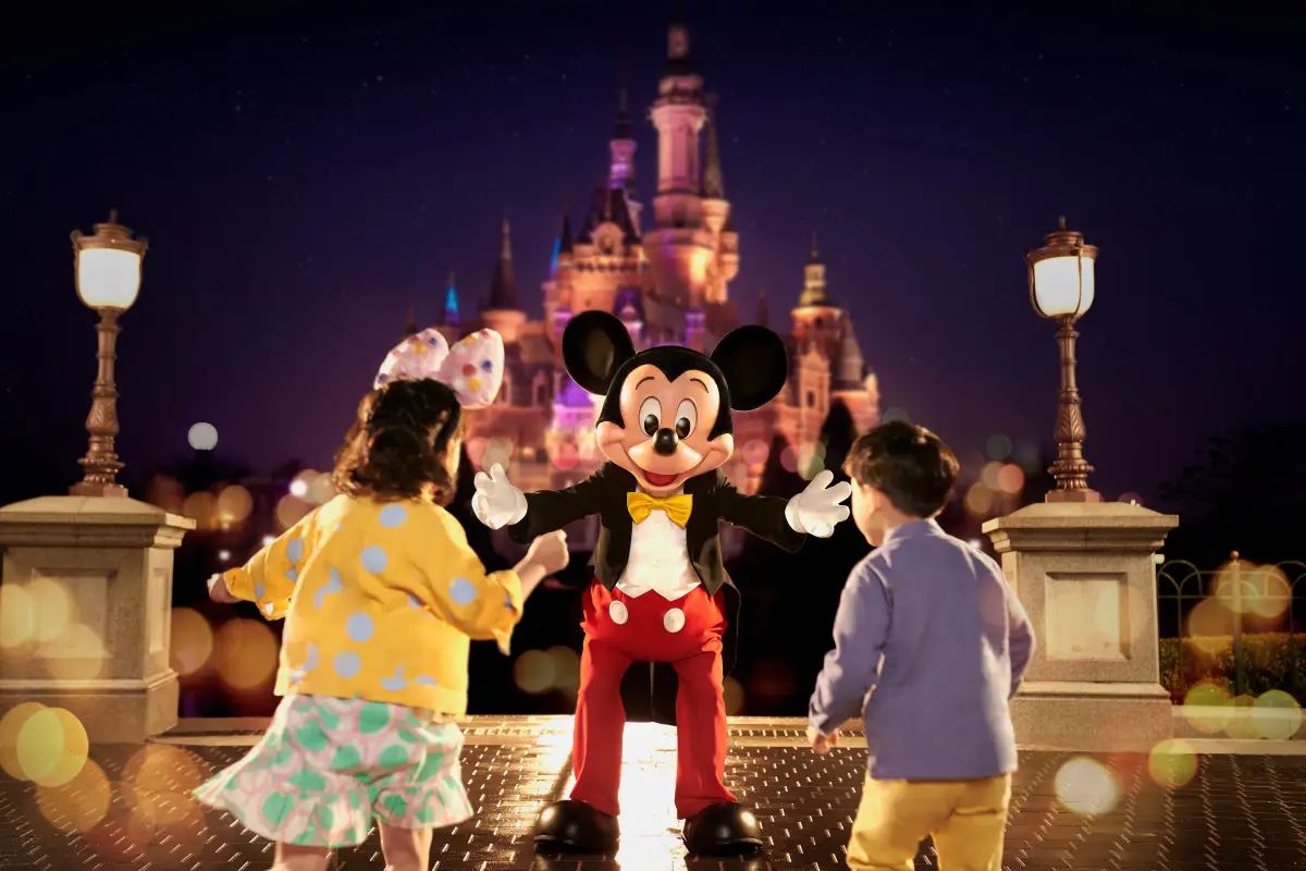 上海ディズニーランドの夜、ライトアップされた城の前でミッキーマウスと子どもたちがふれあう様子。