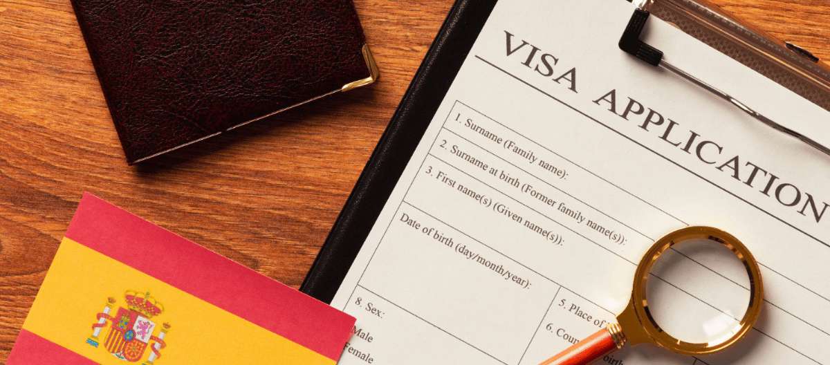 Formulario de solicitud de visa junto a una bandera de España, una lupa y un pasaporte sobre una mesa de madera.
