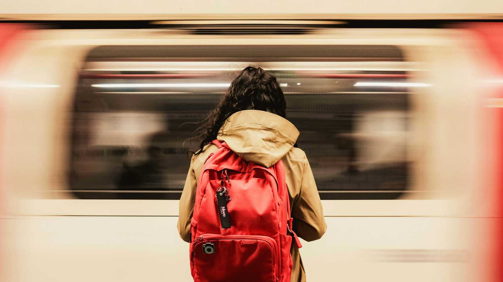 A person seen from behind with a red backpack, waiting as a moving metro train passes by in an underground station