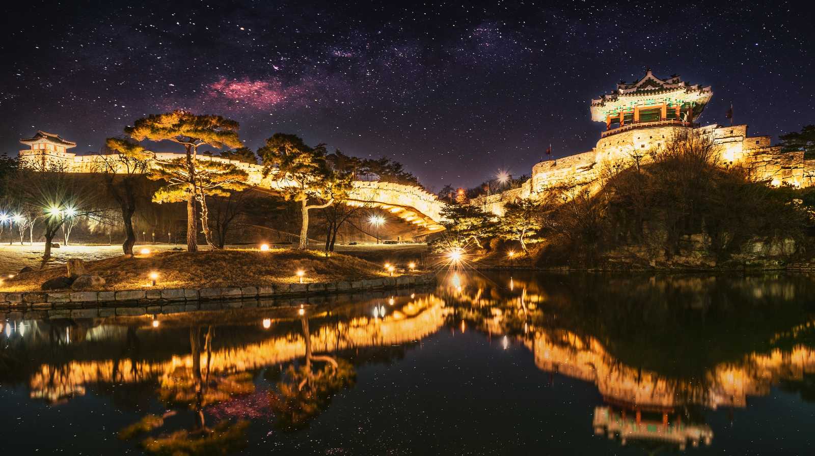 Fortaleza Hwaseong en Suwon iluminada de noche, reflejada en el agua, con un cielo estrellado al fondo