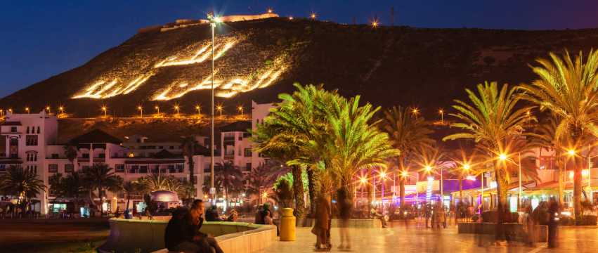 Vista nocturna del paseo marítimo de Agadir con palmeras iluminadas y la colina con letras encendidas.