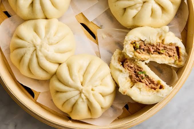 Steamed pork buns, rou baozi, served in a bamboo steamer, with one bun cut open to reveal the filling
