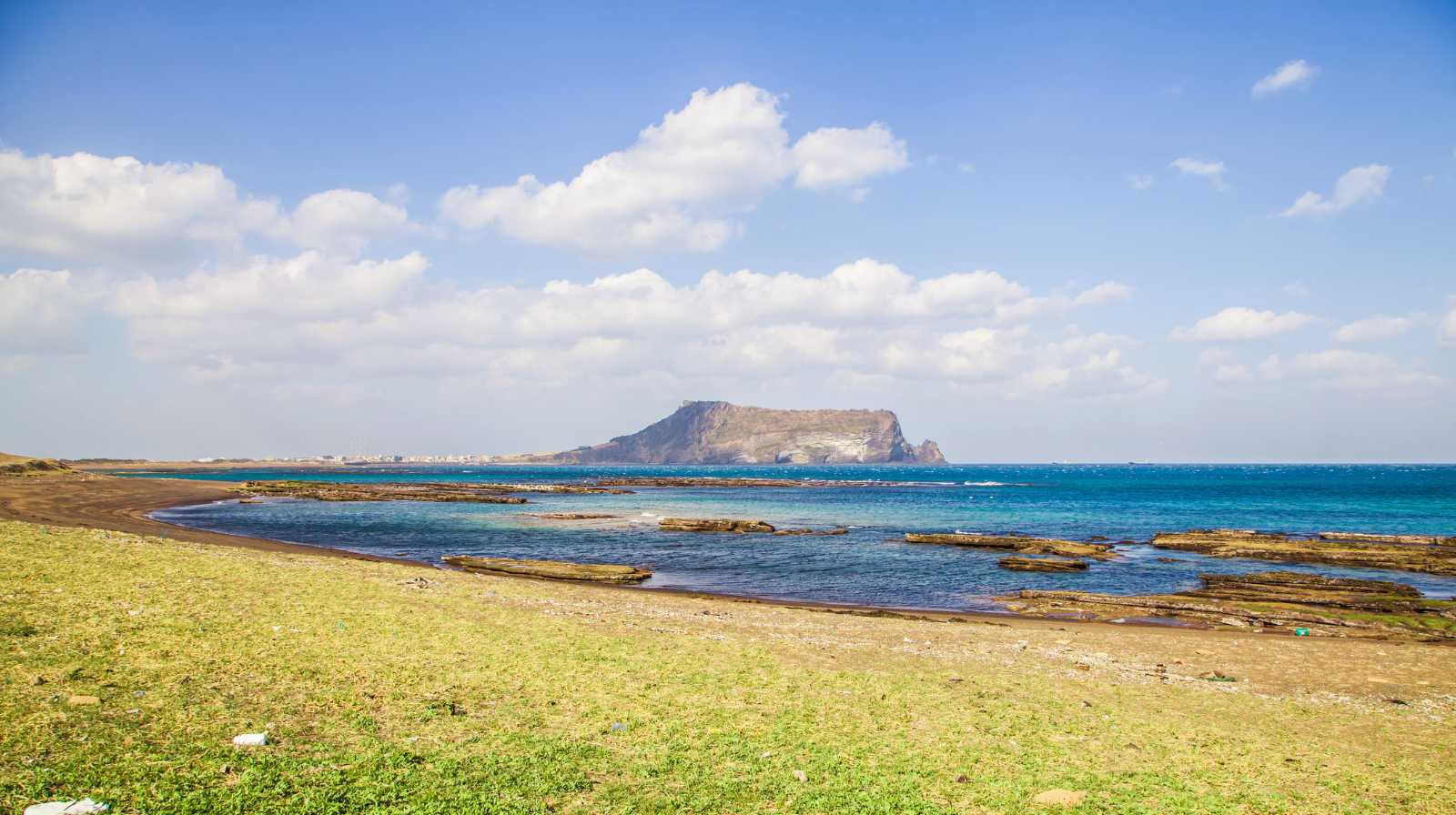 Panorámica costera de Jeju con praderas junto al mar y la montaña Seongsan Ilchulbong elevándose en el horizonte