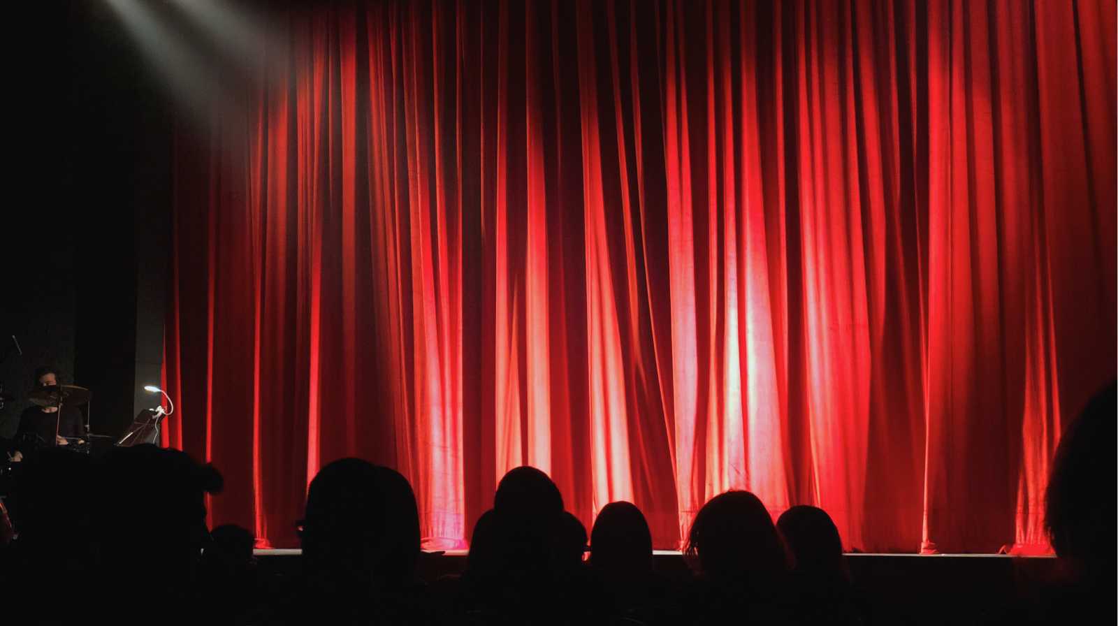 Escenario de teatro con un gran telón rojo iluminado desde arriba, visto desde el público antes de que comience la función