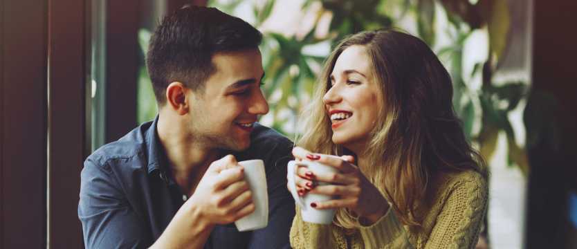 A smiling couple holding coffee cups in a cosy setting by the window