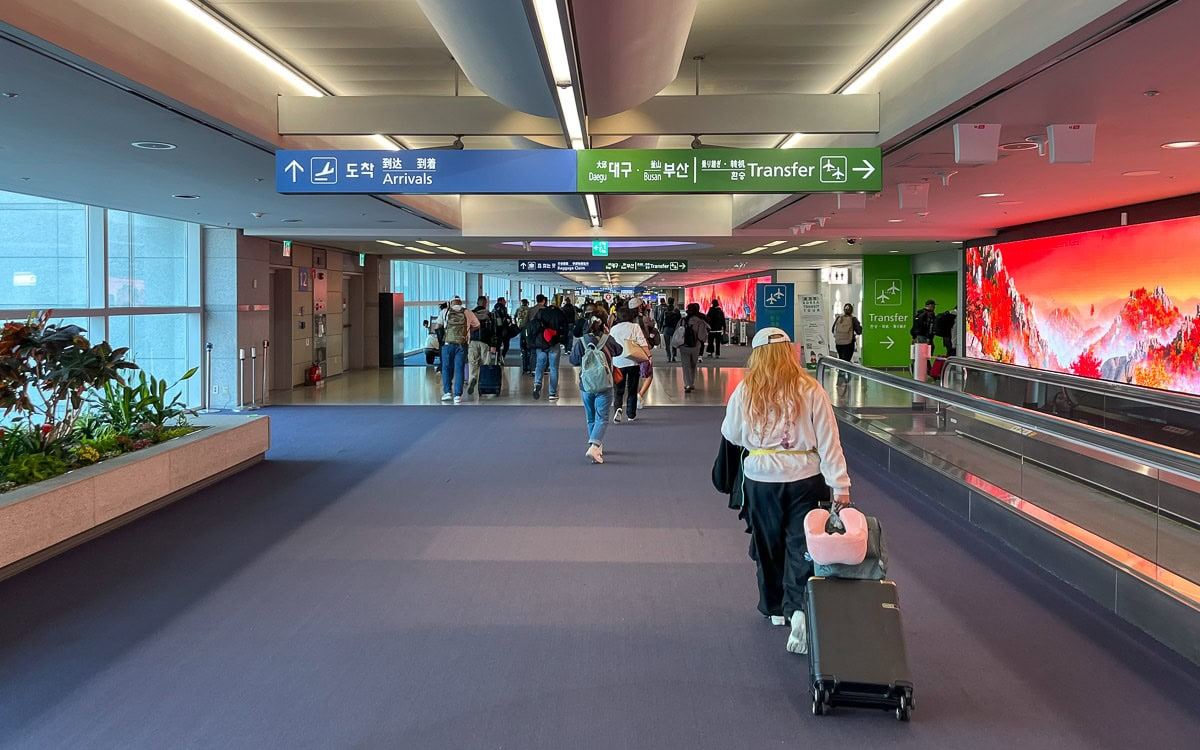Travelers following arrival and transfer signs inside an airport terminal in South Korea.