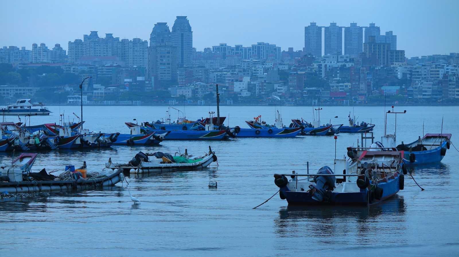 Pequeñas embarcaciones pesqueras amarradas en un puerto de Taipéi con el skyline urbano al fondo en un día nublado.