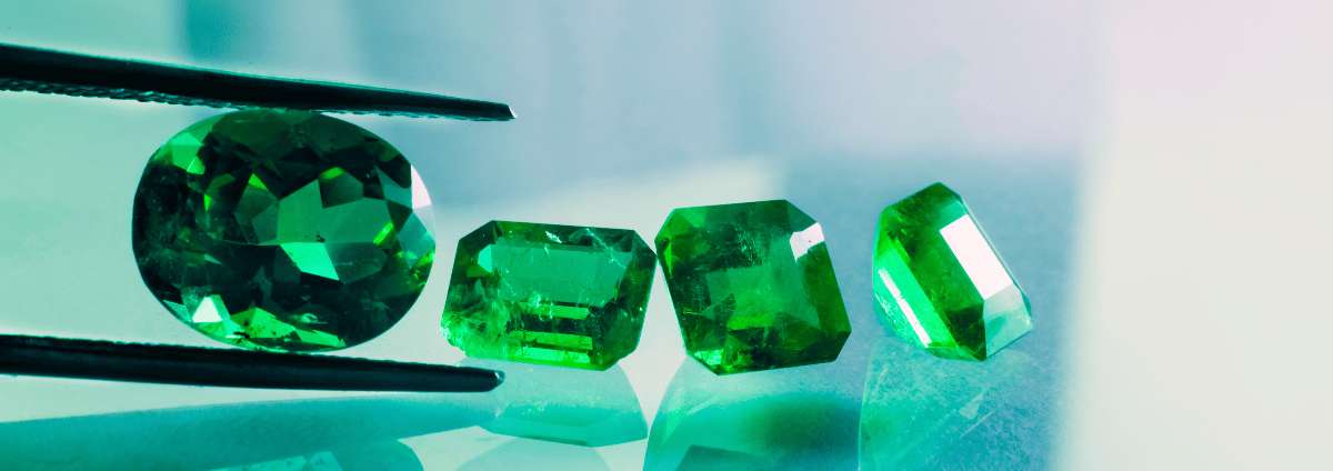 Cut emerald stones held by tweezers on a reflective surface