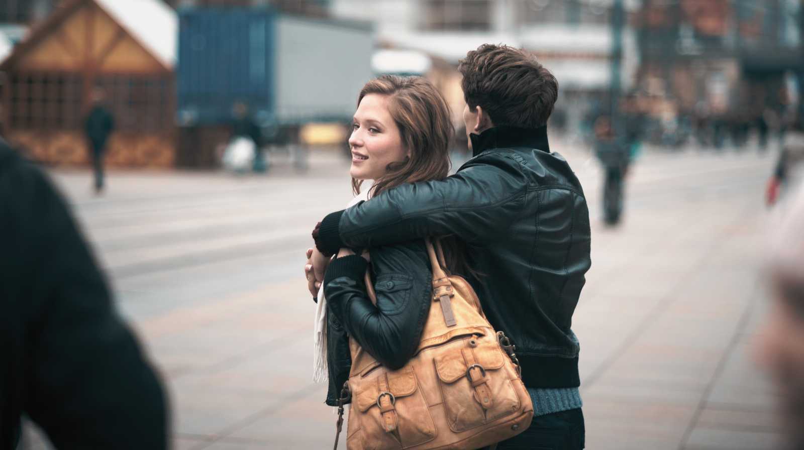 A young couple, arm in arm, strolling through a city, with buildings and people in the background