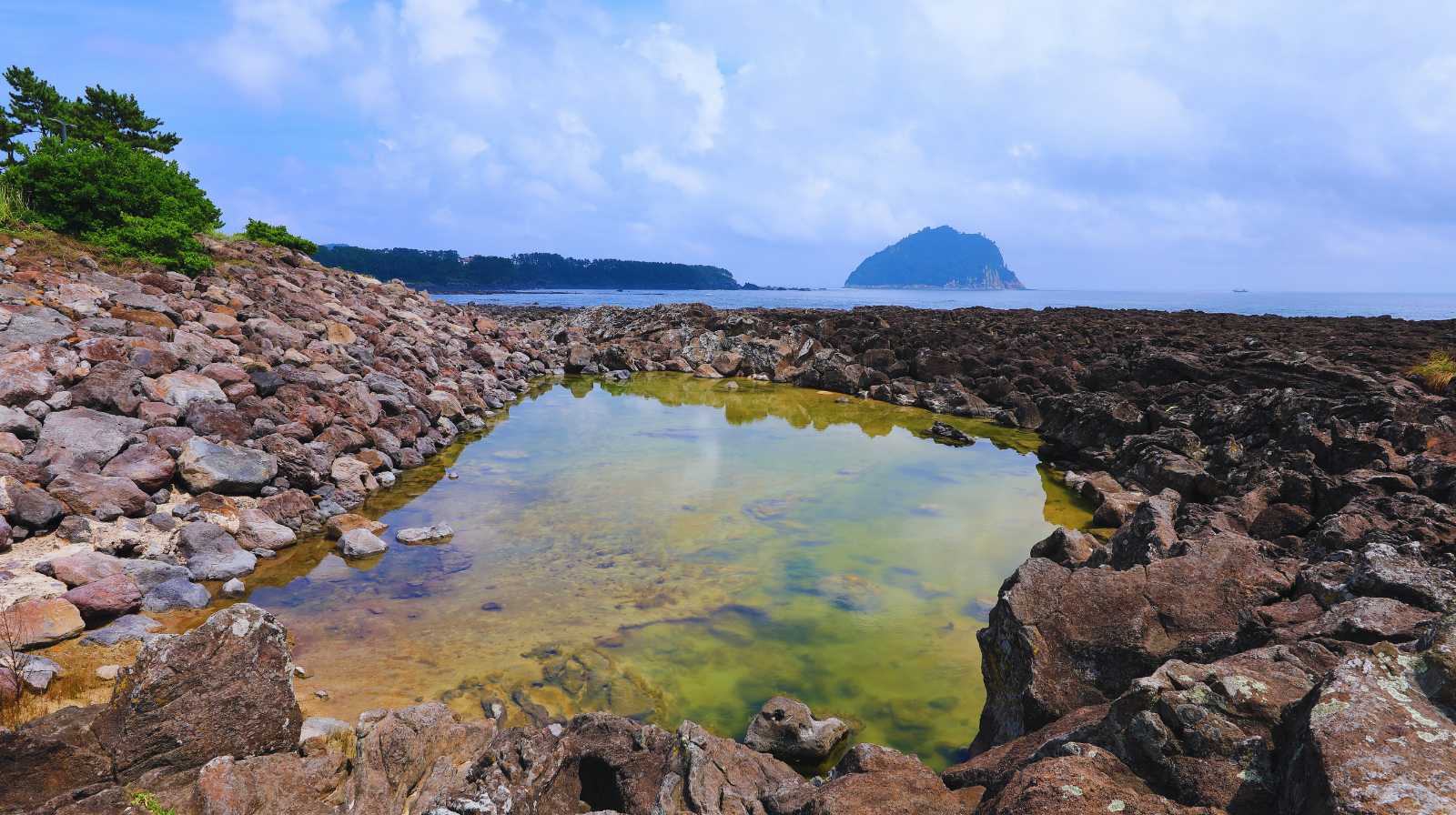 Piscina natural entre rocas volcánicas en la costa de Jeju, con vistas al mar y a una isla montañosa en el horizonte