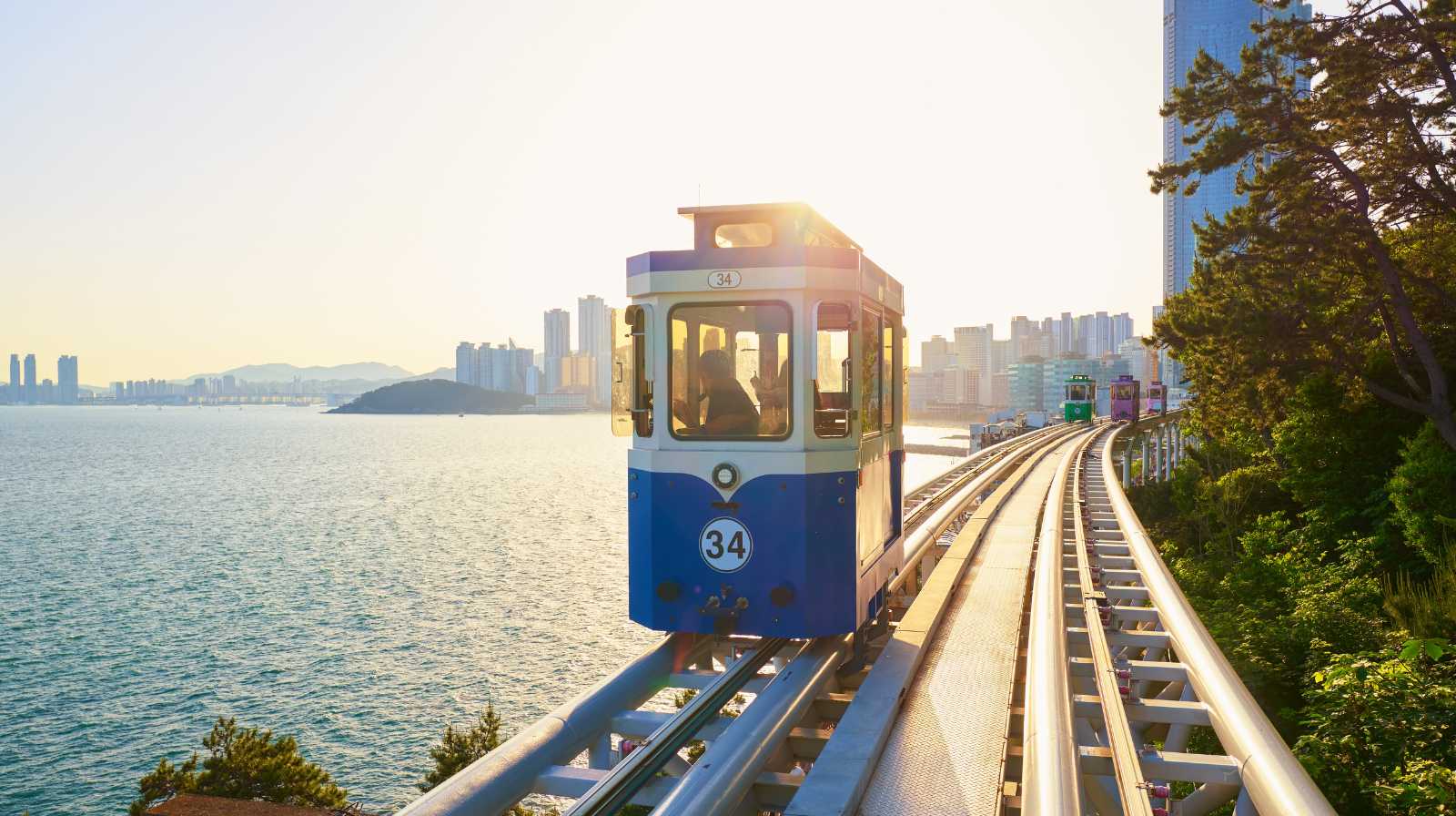 Tren turístico azul avanzando por las vías elevadas junto al mar en Busan durante el atardecer, con los rascacielos de Haeundae al fondo