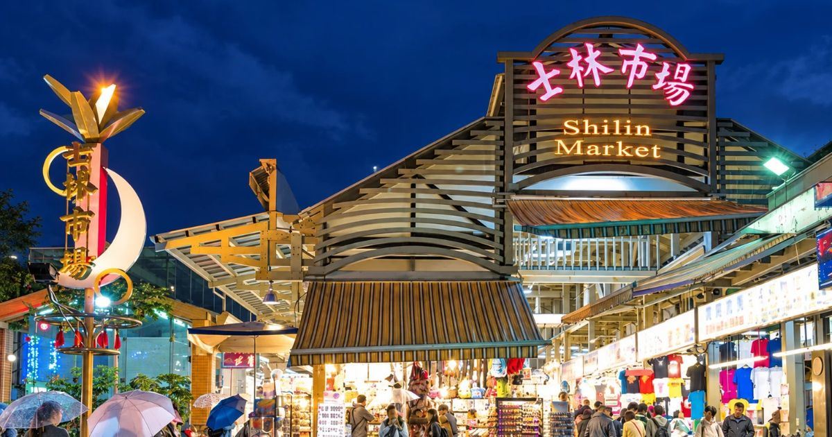 Entrada iluminada del mercado nocturno de Shilin, con puestos comerciales, visitantes y el suelo mojado por la lluvia en Taipéi