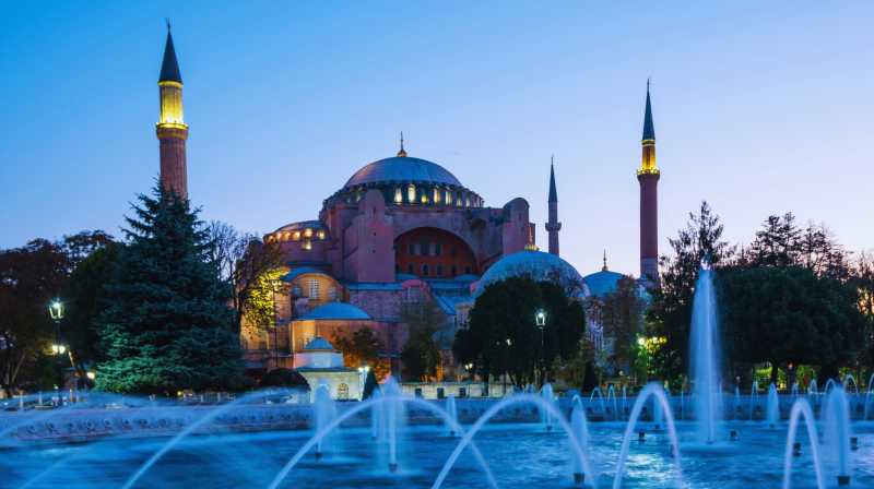 Hagia Sophia illuminated at dusk, with fountains in the foreground and minarets silhouetted against the blue sky