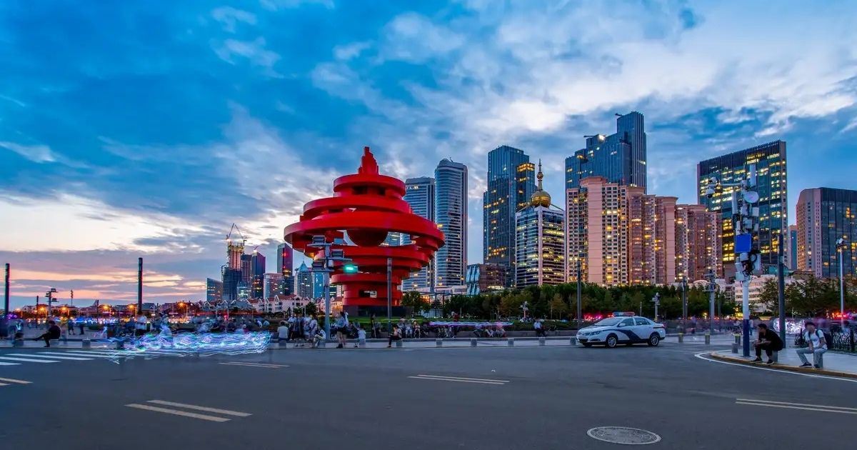 Plaza del 4 de Mayo de Qingdao con la escultura roja May Wind y los rascacielos de la ciudad al atardecer.