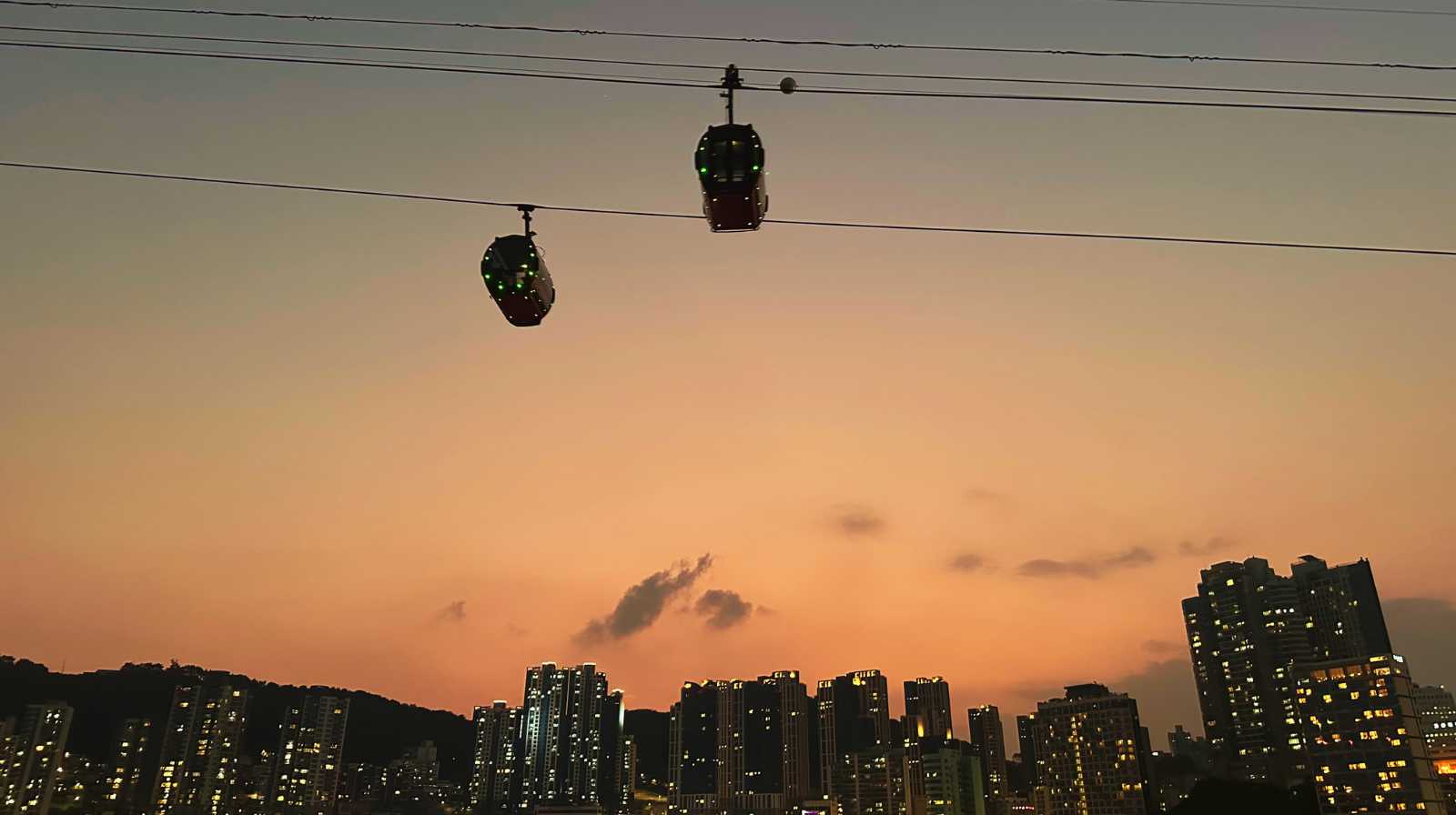 Cabinas de teleférico iluminadas cruzando el cielo durante el atardecer, con la silueta de los edificios de Busan al fondo.