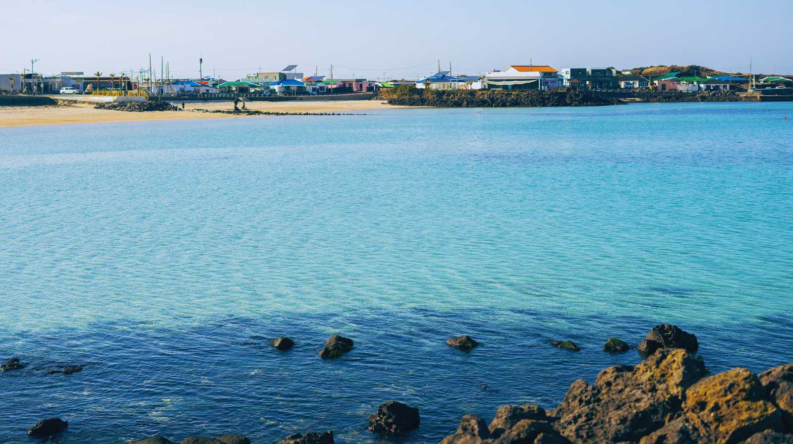 Aguas turquesas junto a una costa volcánica en Jeju, con un pequeño pueblo y playas de arena en la distancia