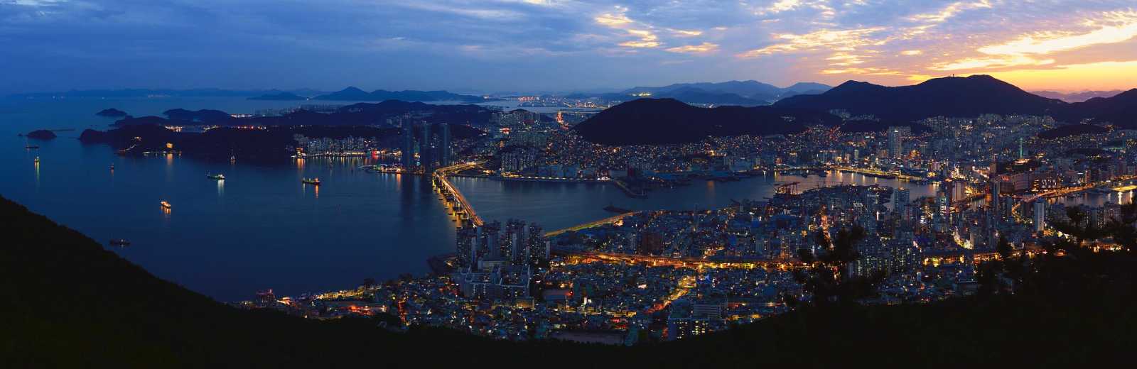 Panorámica nocturna de Jung-gu con puentes iluminados, montañas y la ciudad reflejada sobre el agua