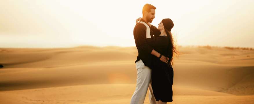 Pareja abrazándose al atardecer en el desierto, con dunas doradas de fondo y luz cálida del sol