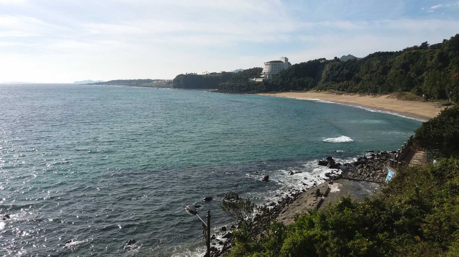 Playa tranquila de Jeju con arena dorada, aguas azules y un hotel rodeado de vegetación en la distancia