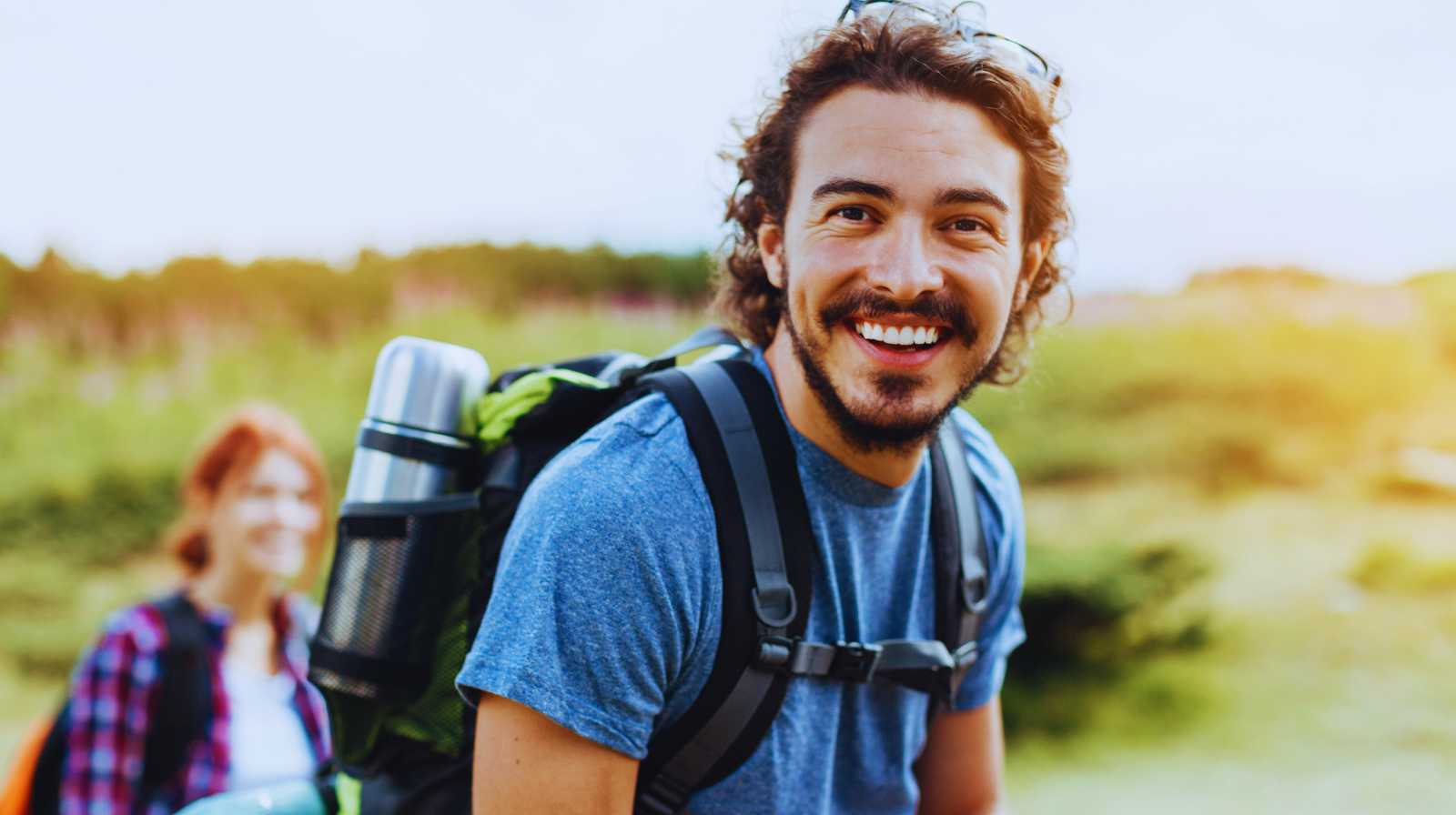 Young traveller with a backpack smiling on an outdoor trail, with another hiker blurred in the background.