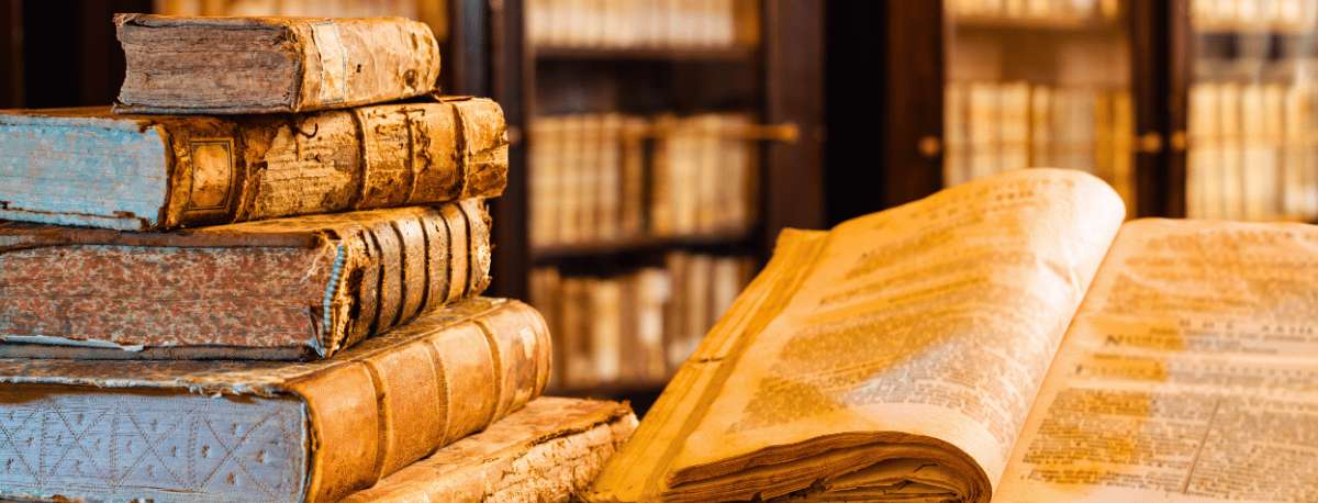 Stack of old books with worn covers next to an open copy in a library