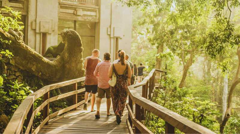 Visitantes caminando por una pasarela de madera entre árboles y esculturas en la selva de Ubud