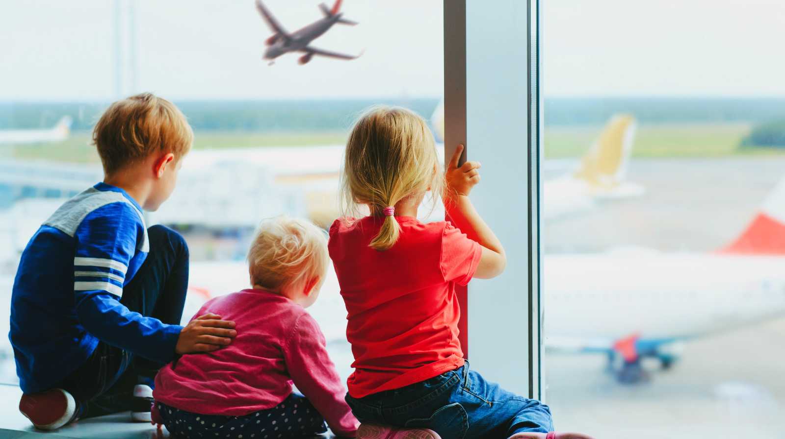 Three children sitting by an airport window watching a plane take off from the runway