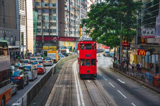 A red double-decker tram on a busy Wan Chai street in Hong Kong, with cars, shops, and pedestrians on both sides